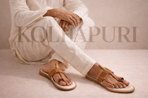 Person wearing white traditional attire with brown sandals on a beige background, featuring the 'Kolhapuri' brand.